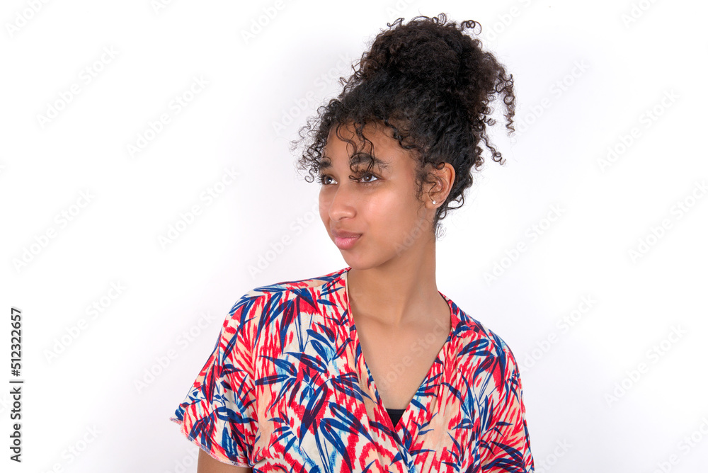 Young African American woman wearing colourful dress over white wall, looks pensively aside, plans actions after university, imagines what to do Thinks over about new project. Young African American woman wearing colourful dress over white wall, looks pensively aside, plans actions after university, imagines what to do Thinks over about new project.