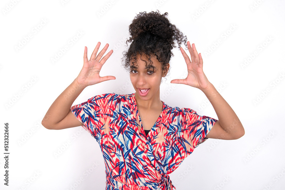 Young African American woman wearing colourful dress over white wall, looks with excitement at camera, keeps hands raised over head, notices something unexpected, reacts on sudden news. Young African American woman wearing colourful dress over white wall, looks with excitement at camera, keeps hands raised over head, notices something unexpected, reacts on sudden news.