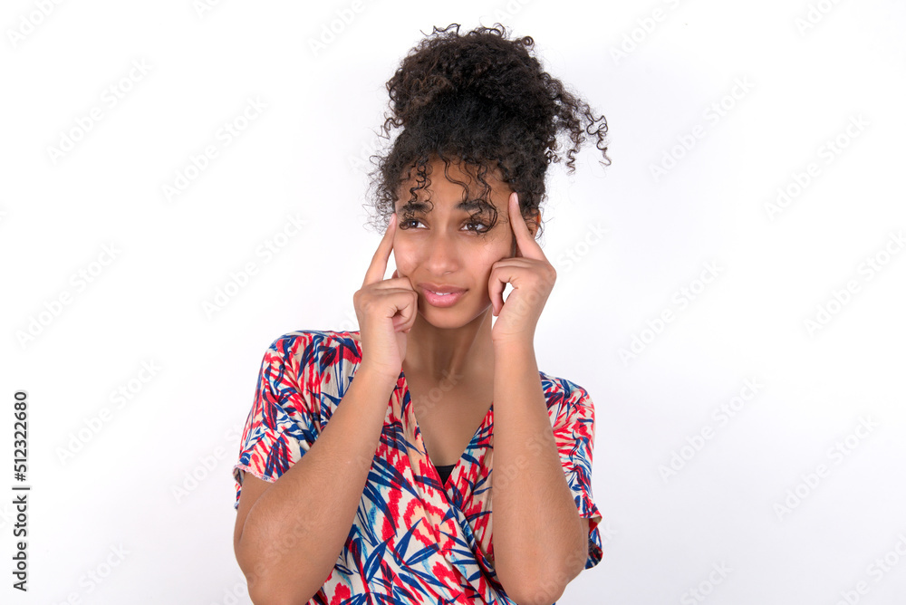 Young African American woman wearing colourful dress over white wall with thoughtful expression, looks away, keeps hand near face, thinks about something pleasant. Young African American woman wearing colourful dress over white wall with thoughtful expression, looks away, keeps hand near face, thinks about something pleasant.