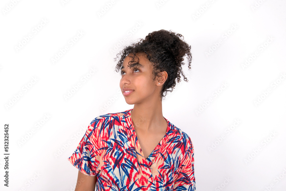 Young African American woman wearing colourful dress over white wall looking up as he sees something strange. Young African American woman wearing colourful dress over white wall looking up as he sees something strange.