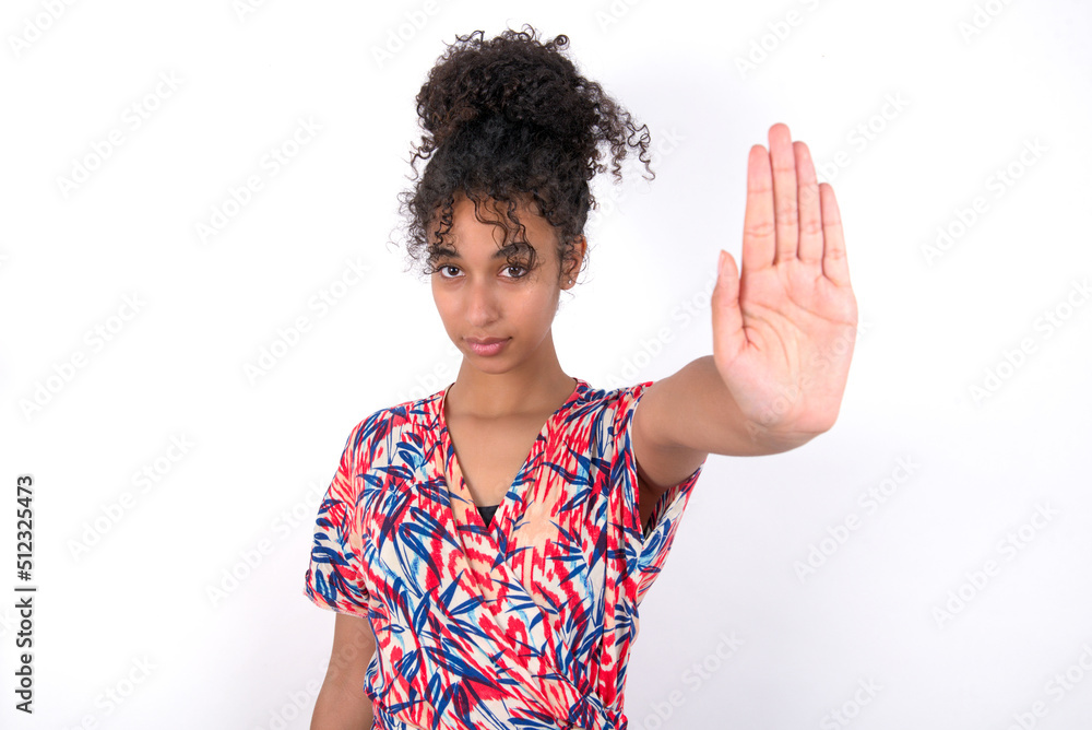 Young African American woman wearing colourful dress over white wall doing stop gesture with palm of the hand. Warning expression with negative and serious gesture on the face. Young African American woman wearing colourful dress over white wall doing stop gesture with palm of the hand. Warning expression with negative and serious gesture on the face.