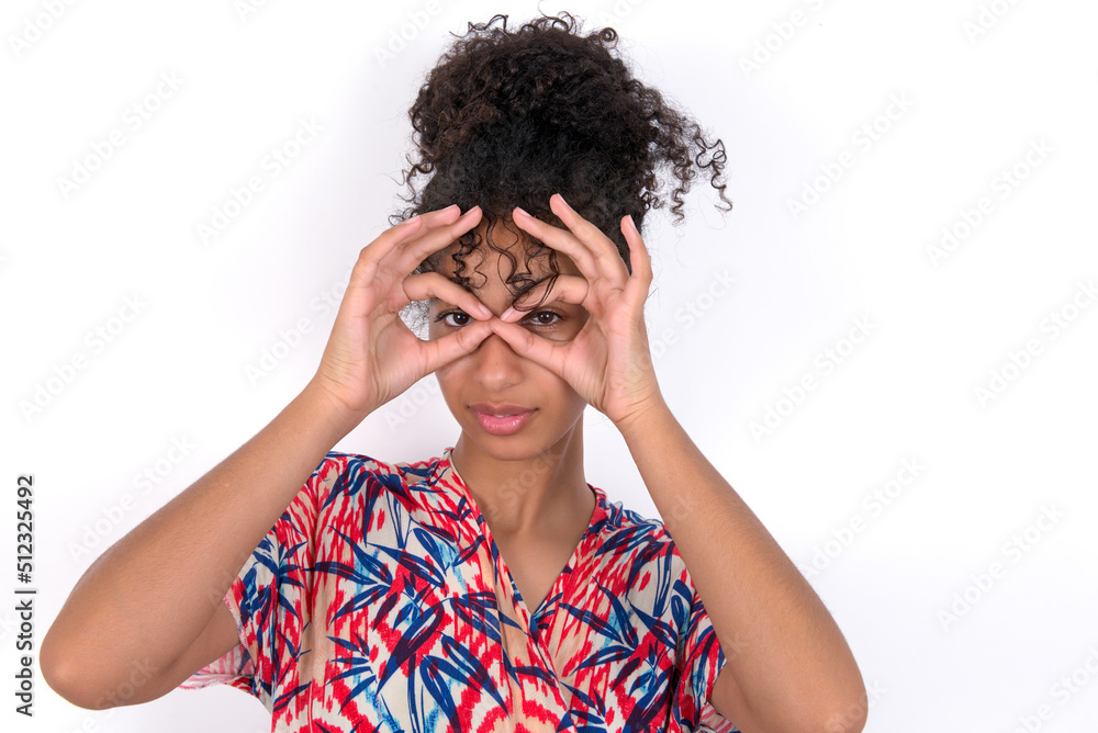 Playful excited Young African American woman wearing colourful dress over white wall showing Ok sign with both hands on eyes, pretending to wear spectacles. Playful excited Young African American woman wearing colourful dress over white wall showing Ok sign with both hands on eyes, pretending to wear spectacles.