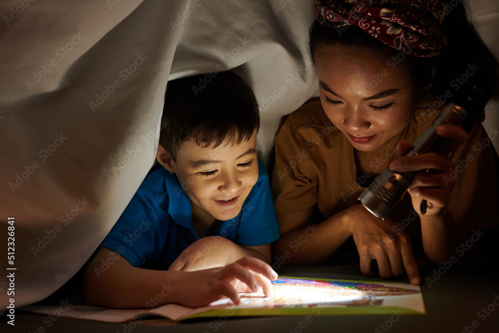 Mother shining flashlight on book page when son reading scary story - obrazy, fototapety, plakaty 