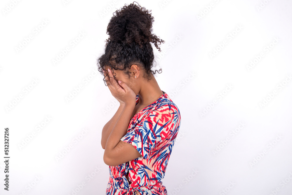 Sad Young African American woman wearing colourful dress over white wall covering face with hands and crying. Sad Young African American woman wearing colourful dress over white wall covering face with hands and crying.