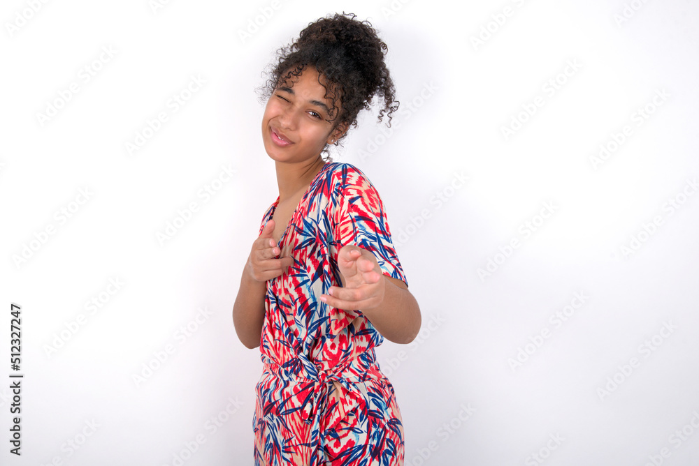 Hey you, bang. Joyful and charismatic good-looking Young African American woman wearing colourful dress over white wall winking and pointing with finger pistols at camera happily and cheeky. Hey you, bang. Joyful and charismatic good-looking Young African American woman wearing colourful dress over white wall winking and pointing with finger pistols at camera happily and cheeky.