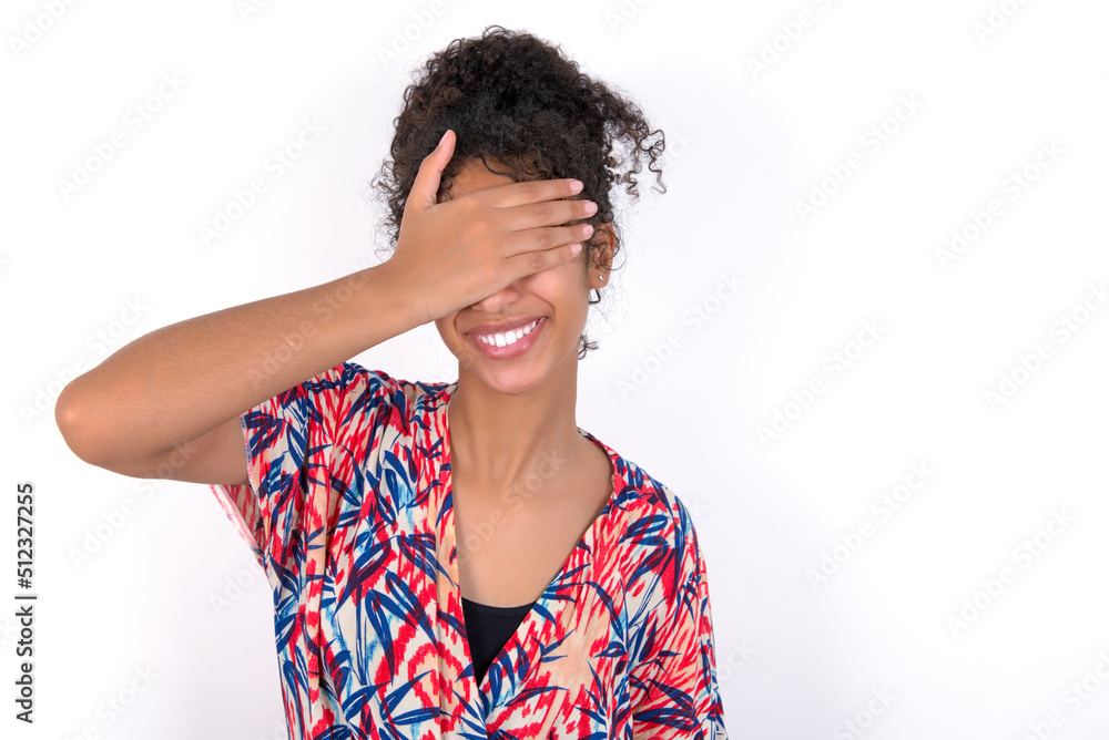 Happy Young African American woman wearing colourful dress over white wall closing eyes with hand going to see surprise prepared by friend standing and smiling in anticipation for something wonderful. Happy Young African American woman wearing colourful dress over white wall closing eyes with hand going to see surprise prepared by friend standing and smiling in anticipation for something wonderful.