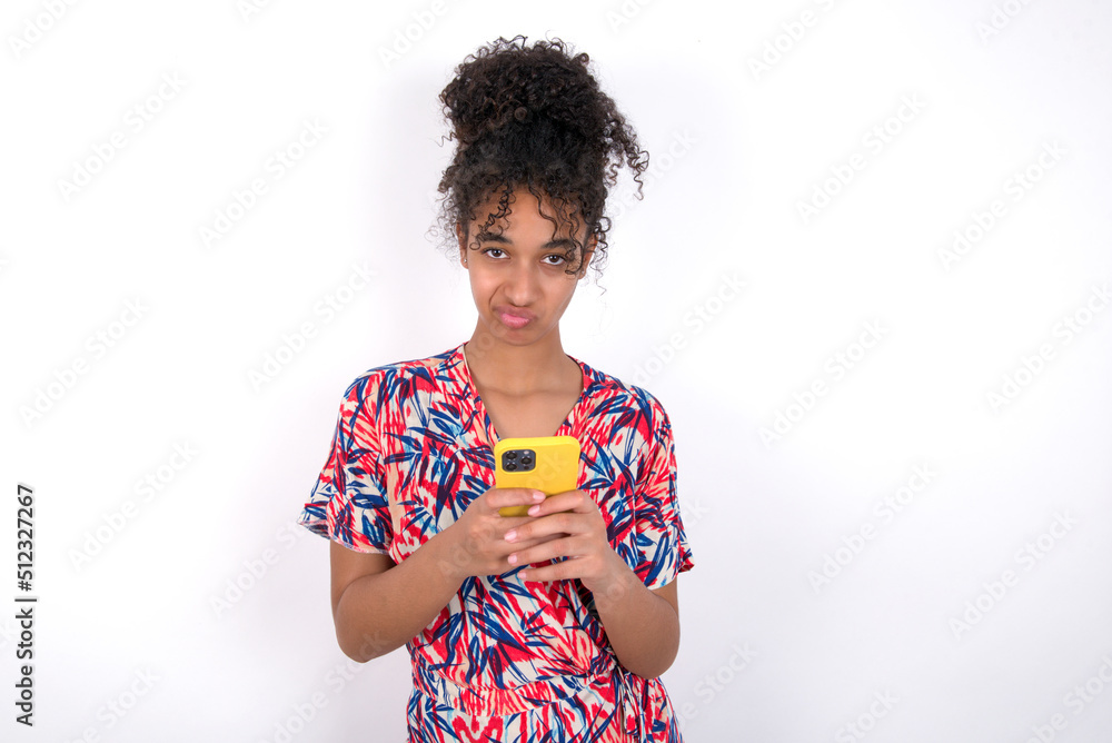 Portrait of a confused Young African American woman wearing colourful dress over white wall holding mobile phone and shrugging shoulders and frowning face.