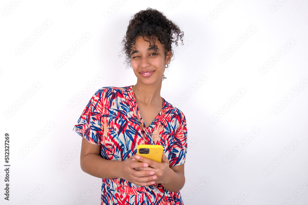 Pleased Young African American woman wearing colourful dress over white wall using self phone and looking and winking at the camera. Flirt and coquettish concept. Pleased Young African American woman wearing colourful dress over white wall using self phone and looking and winking at the camera. Flirt and coquettish concept.