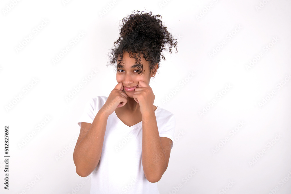 Pleased Young beautiful girl with afro hairstyle wearing white t-shirt over white wall with closed eyes keeps hands near cheeks and smiles tenderly imagines something very pleasant