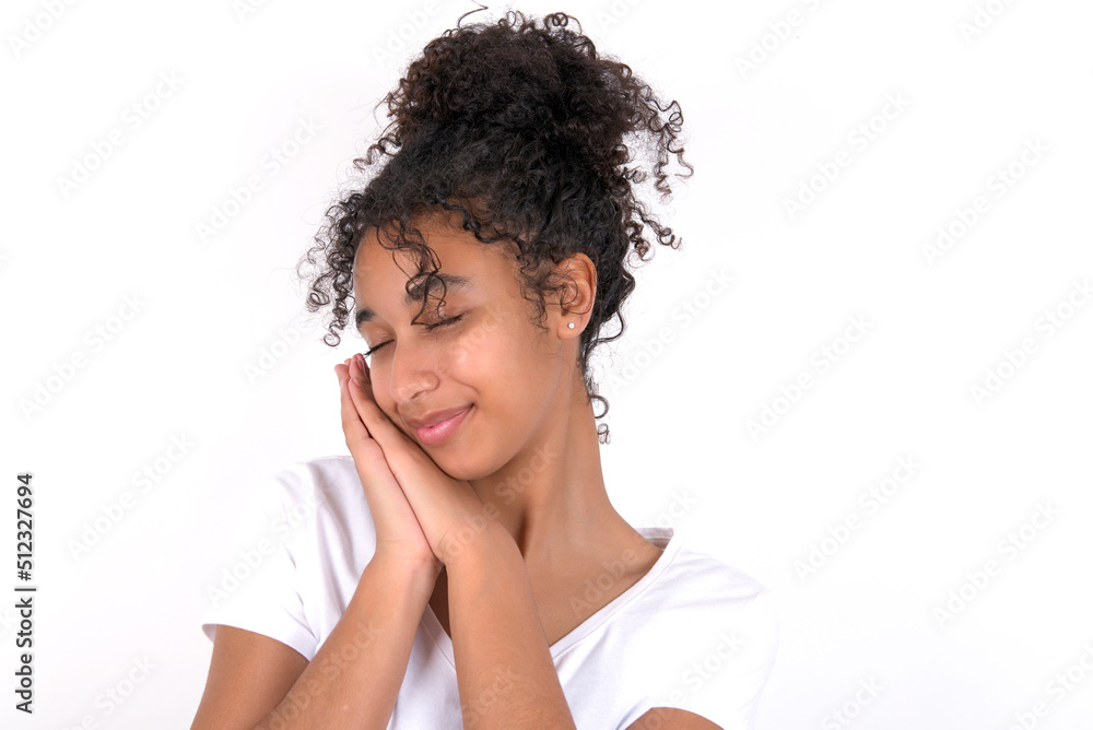 Young beautiful girl with afro hairstyle wearing white t-shirt over white wall leans on pressed palms closes eyes and has pleasant smile dreams about something Young beautiful girl with afro hairstyle wearing white t-shirt over white wall leans on pressed palms closes eyes and has pleasant smile dreams about something