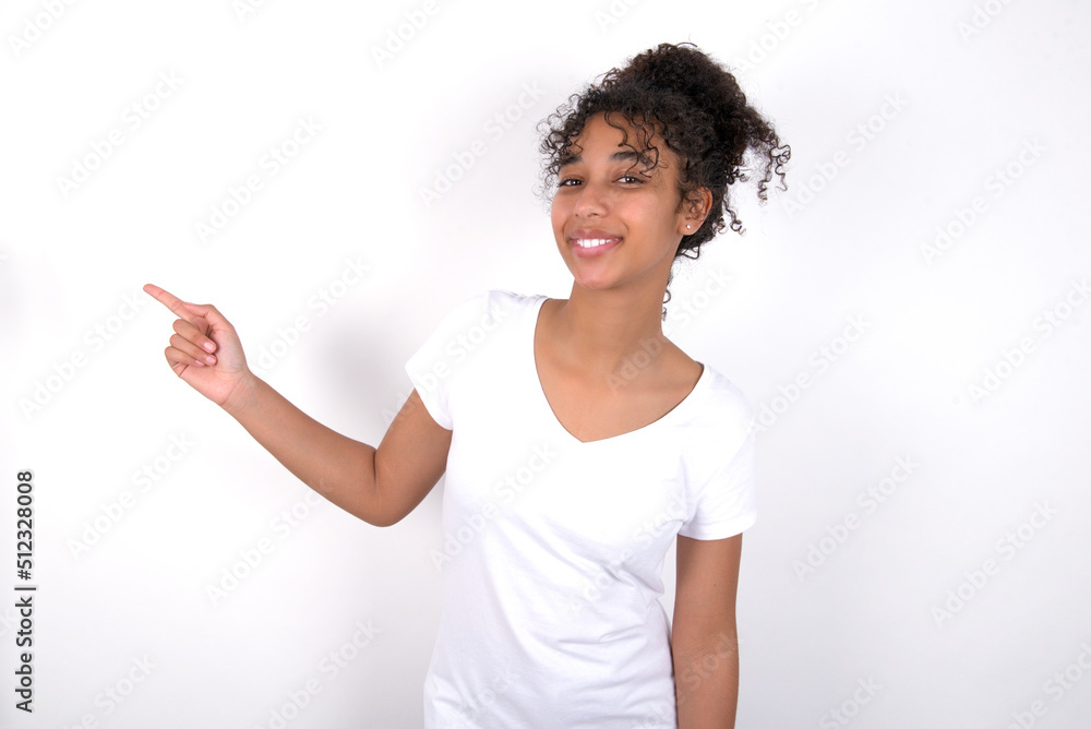 Young beautiful girl with afro hairstyle wearing white t-shirt over white wall laughs happily points away on blank space demonstrates shopping discount offer, excited by good news or unexpected sale. Young beautiful girl with afro hairstyle wearing white t-shirt over white wall laughs happily points away on blank space demonstrates shopping discount offer, excited by good news or unexpected sale.