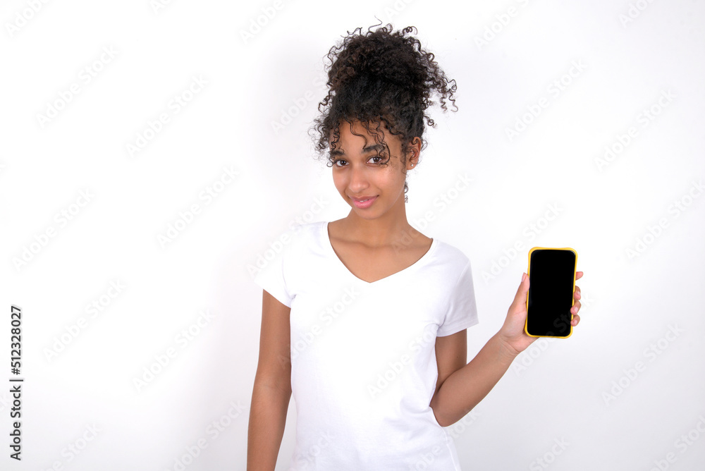 Young beautiful girl with afro hairstyle wearing white t-shirt over white wall holds new mobile phone and looks mysterious aside shows blank display of modern cellular Young beautiful girl with afro hairstyle wearing white t-shirt over white wall holds new mobile phone and looks mysterious aside shows blank display of modern cellular