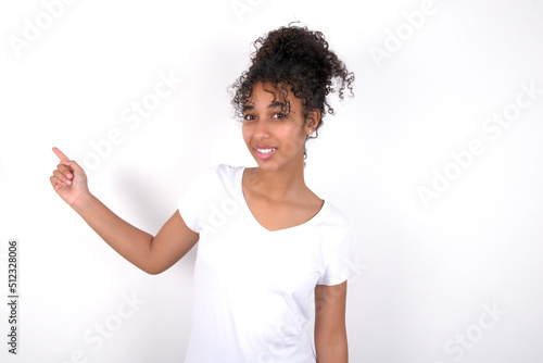 Young beautiful girl with afro hairstyle wearing white t-shirt over white wall points aside on copy blank space. People promotion and advertising concept