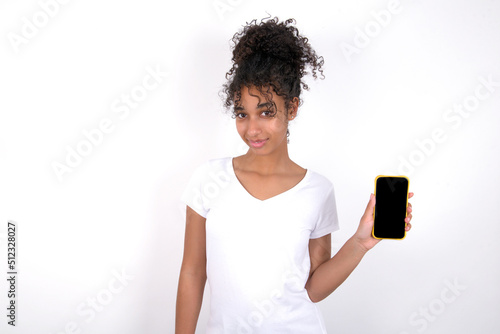 Young beautiful girl with afro hairstyle wearing white t-shirt over white wall holds new mobile phone and looks mysterious aside shows blank display of modern cellular