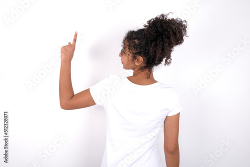 Young beautiful girl with afro hairstyle wearing white t-shirt over white background pointing to object on copy space, rear view. Turn your back
