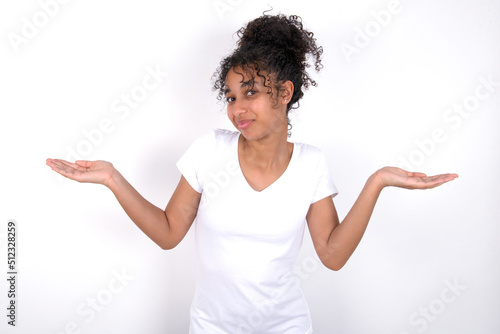 Careless attractive Young beautiful girl with afro hairstyle wearing white t-shirt over white background shrugging shoulders, oops.