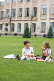 Schoolgirl eating sandwich near asian friend and lunchboxes on lawn in park.