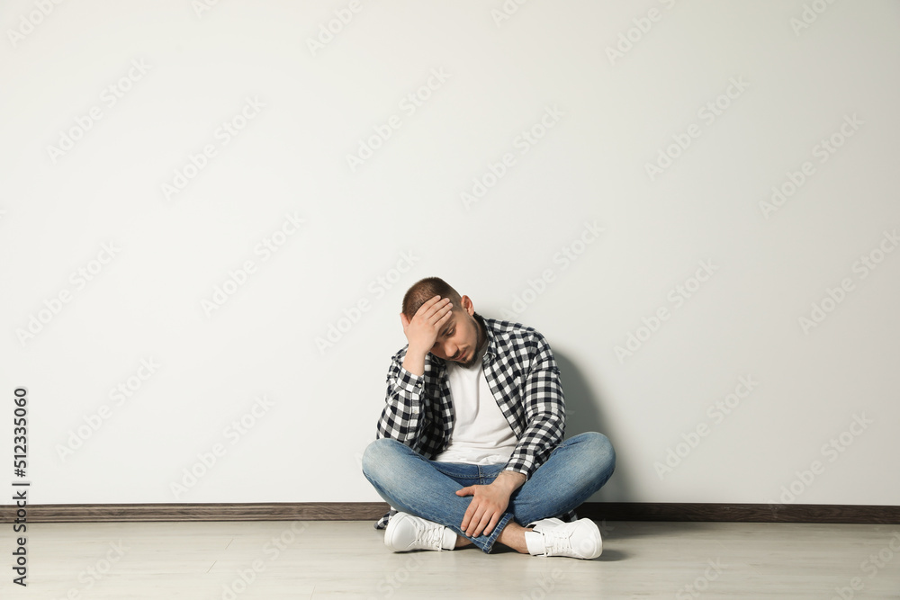 Sad young man sitting on floor near white wall indoors, space for text Sad young man sitting on floor near white wall indoors, space for text