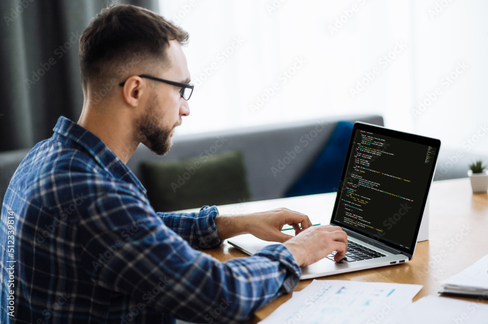 Developing coding technologies, IT concept. Side view at male IT-specialist uses laptop with programming code on the screen. Smart male programmer working at home with coding technology, writing codes Developing coding technologies, IT concept. Side view at male IT-specialist uses laptop with programming code on the screen. Smart male programmer working at home with coding technology, writing codes