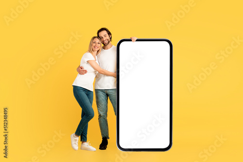 Cheerful millennial caucasian man and female in white t-shirts hugging, stand near huge phone with blank screen