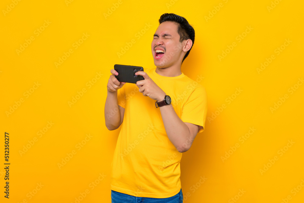 Excited young Asian man in casual t-shirt playing online game on mobile phone isolated on yellow background. People lifestyle concept Excited young Asian man in casual t-shirt playing online game on mobile phone isolated on yellow background. People lifestyle concept