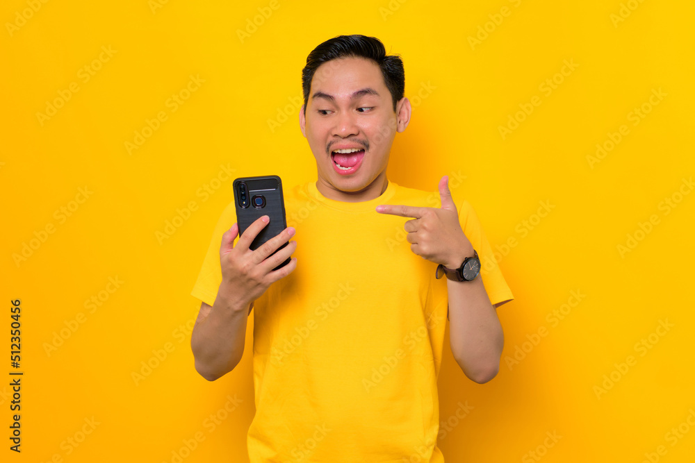 Excited young Asian man in casual t-shirt pointing fingers at mobile phone, reacting to new app isolated on yellow background. People lifestyle concept Excited young Asian man in casual t-shirt pointing fingers at mobile phone, reacting to new app isolated on yellow background. People lifestyle concept