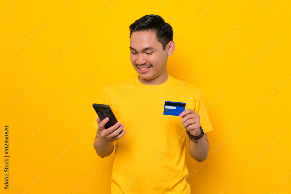 Smiling young Asian man in casual t-shirt holding mobile phone and credit card isolated on yellow background. People lifestyle concept Smiling young Asian man in casual t-shirt holding mobile phone and credit card isolated on yellow background. People lifestyle concept