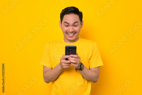 Smiling young Asian man in casual t-shirt using mobile phone and looking at gadget screen isolated on yellow background. People lifestyle concept © Sewupari Studio