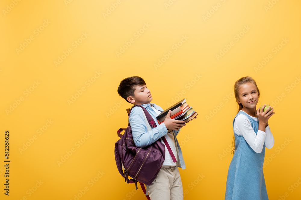 Asian schoolboy with backpack holding books near friend with apple isolated on yellow.