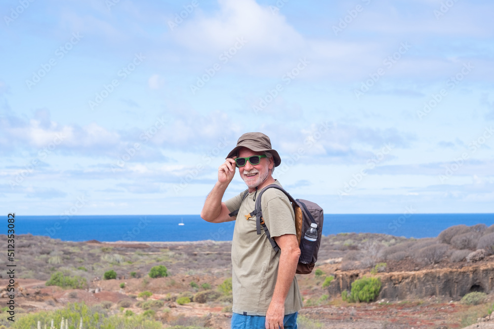 Happy active senior man with backpack and hat while hiking outdoors looking at camera smiling. Elderly man enjoying retirement and healthy lifestyle. Blue sea and sky in the background Happy active senior man with backpack and hat while hiking outdoors looking at camera smiling. Elderly man enjoying retirement and healthy lifestyle. Blue sea and sky in the background