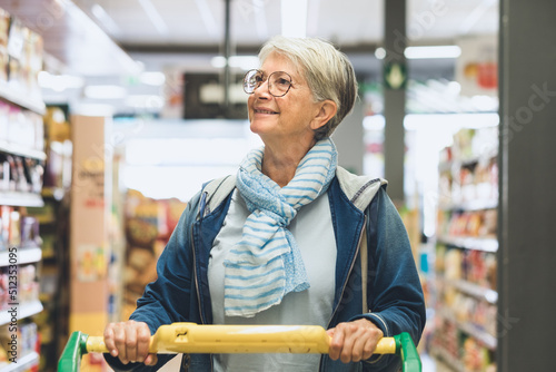 Senior caucasian woman pushing a shopping trolley in the supermarket looking aat the products on display, customer attentive to shopping costs photo