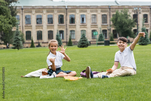 Positive schoolgirl holding sandwich and waving hand at camera near asian friend on lawn in park.