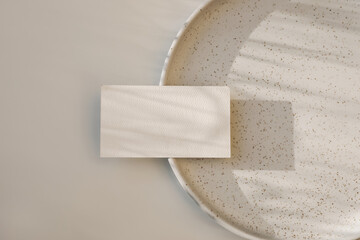 Modern neutral branding stationery. Closeup of textured blank business card, invitation mockup on dotted, speckled ceramic plate in sunlight. Beige table background. Flat lay, top view. No people.