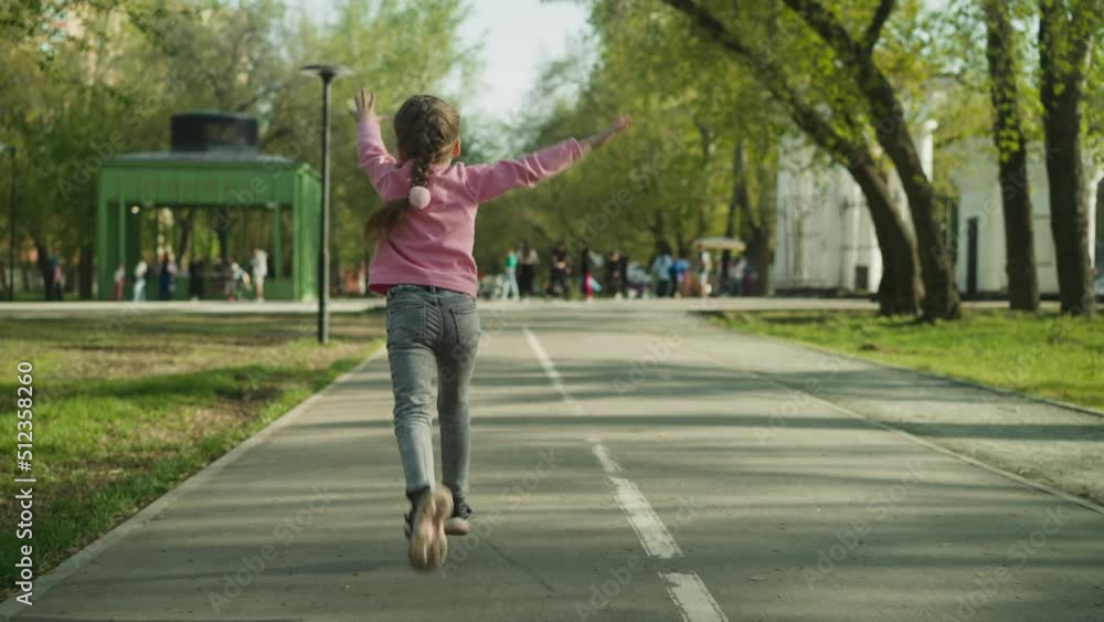 Happy little girl with plait runs waving hands along road in spring city park backside view. Joyful child enjoys nice weather in public garden slow motion Happy little girl with plait runs waving hands along road in spring city park backside view. Joyful child enjoys nice weather in public garden slow motion