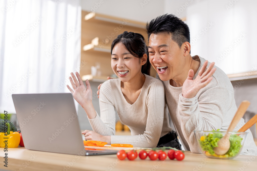Happy asian spouses having video call while cooking at kitchen