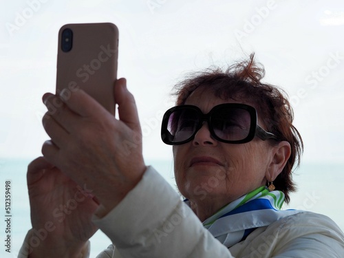 Obraz na plátně An elderly woman uses a cell phone, takes a selfie from the balcony of the sanatorium against the backdrop of the sea