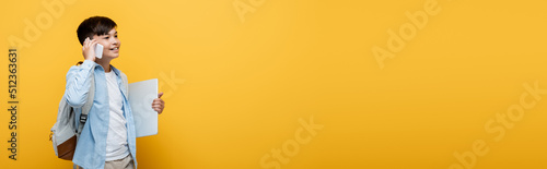 Positive asian schoolboy talking on smartphone and holding laptop isolated on yellow, banner.