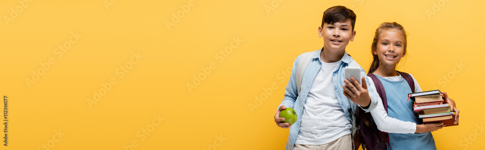 Preteen interracial schoolkids holding books and mobile phone isolated on yellow, banner. Preteen interracial schoolkids holding books and mobile phone isolated on yellow, banner.