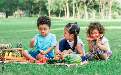 Three little mixed race kids consist of African and caucasian boys and girls smiling with happiness, fun amusement, playing, sitting for picnic and eating piece of watermelon fruit in outdoor garden.