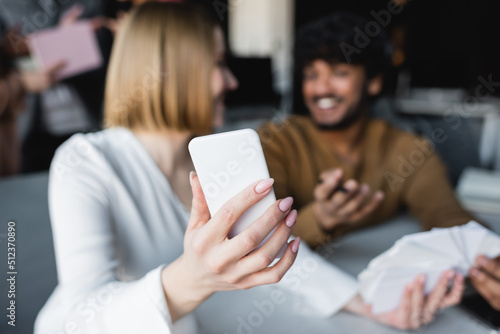 selective focus of smartphone in hand of woman near indian man smiling on blurred background.