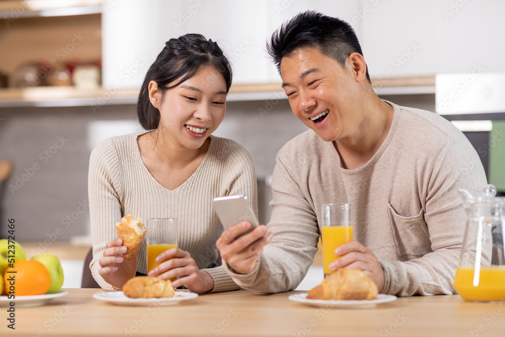 Happy asian couple using cell phone while having breakfast