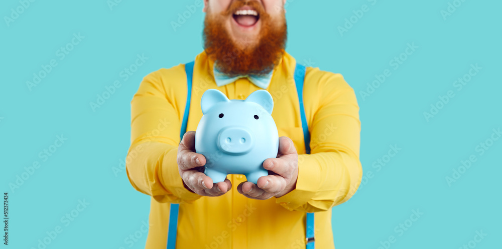 Piggy bank in form of blue pig in hands of joyful contented unrecognizable bearded fat man. Man in yellow shirt and suspenders holding piggy bank on light blue background. Close up. Cropped image. Piggy bank in form of blue pig in hands of joyful contented unrecognizable bearded fat man. Man in yellow shirt and suspenders holding piggy bank on light blue background. Close up. Cropped image.