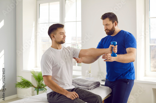 Professional physiotherapist controls his patient who is doing arm muscle rehabilitation exercises. Serious young man doing physical exercises with dumbbells at modern physiotherapy clinic or center