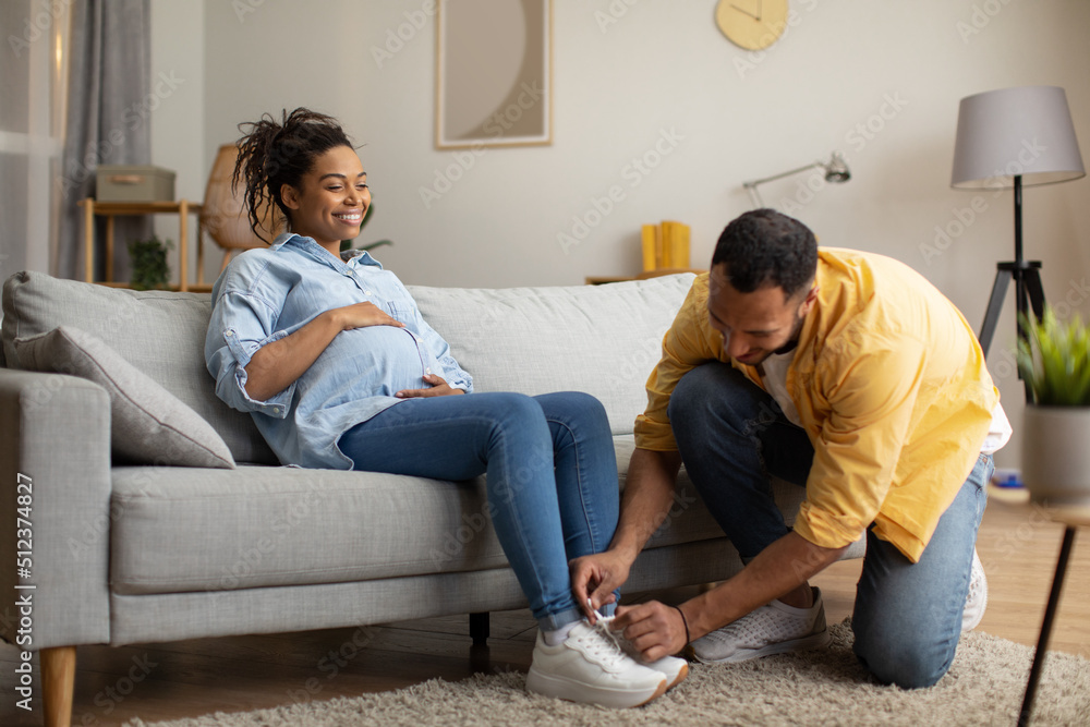 Fototapeta premium Loving African American Husband Helping Pregnant Wife Lacing Shoes Indoors