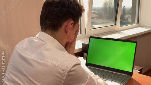 A frustrated Caucasian man sits at a desk in front of a computer with a green screen in the office and rubs his face with his hands. Workaholic, hard work, career ladder, earning money. High quality