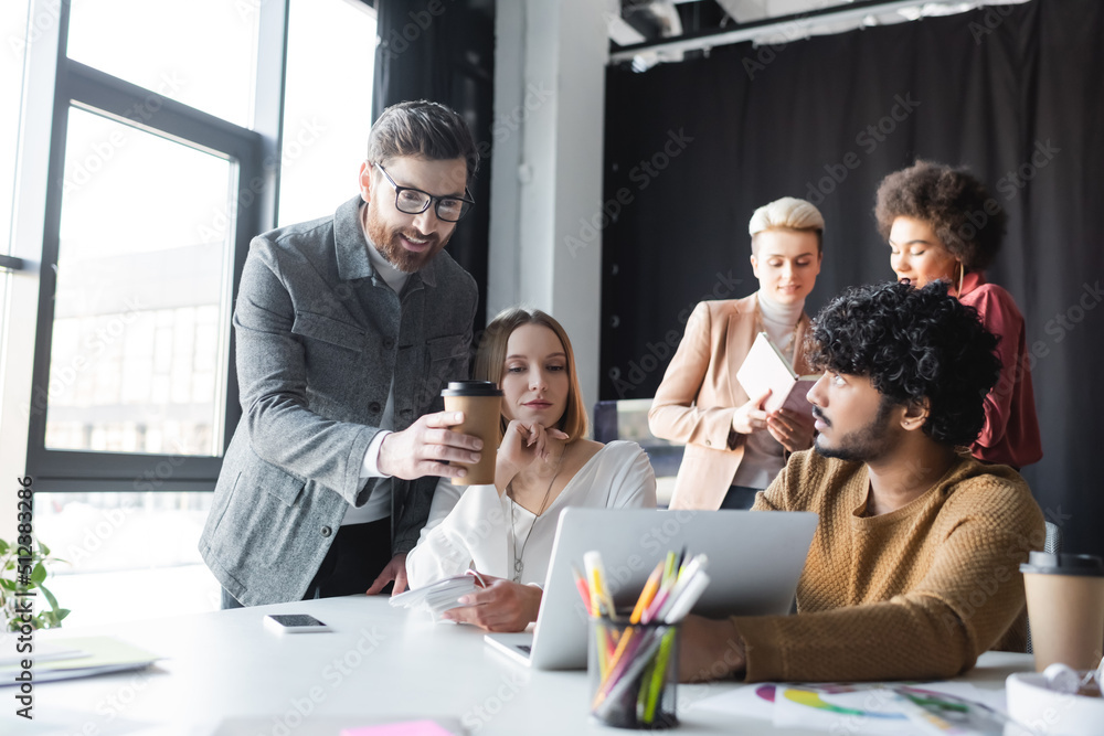 smiling man with paper cup pointing at laptop near interracial advertising managers. smiling man with paper cup pointing at laptop near interracial advertising managers.