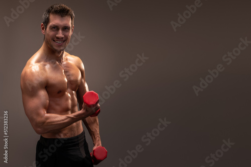 Smiling athletic man holding red dumbbell in hand and looking at the camera, recommending you to start pumping up muscles. Indoor studio shot isolated on brown background