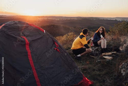Two Young Travelers Man and Woman Sitting Near the Campfire and Tent During Sunset Over Top of Hill