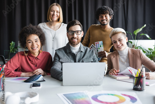 happy multiethnic advertising designers looking at camera near laptop and color samples.