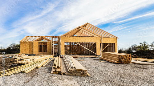 New house under construction, front view from garage door entry with clouds blue sky. Framing of home exterior structure with pile of lumber and siding plamk leftover. Real estate business industry.